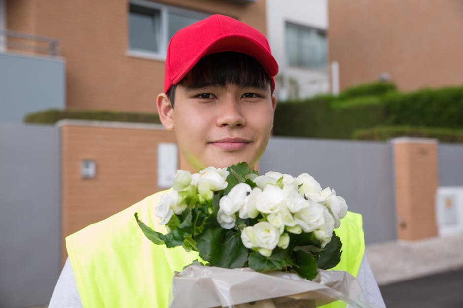 A young male flower delivery courier wearing a red cap and a neon yellow safety vest is standing outdoors in front of modern residential buildings. He is holding a bouquet of fresh white flowers, incl