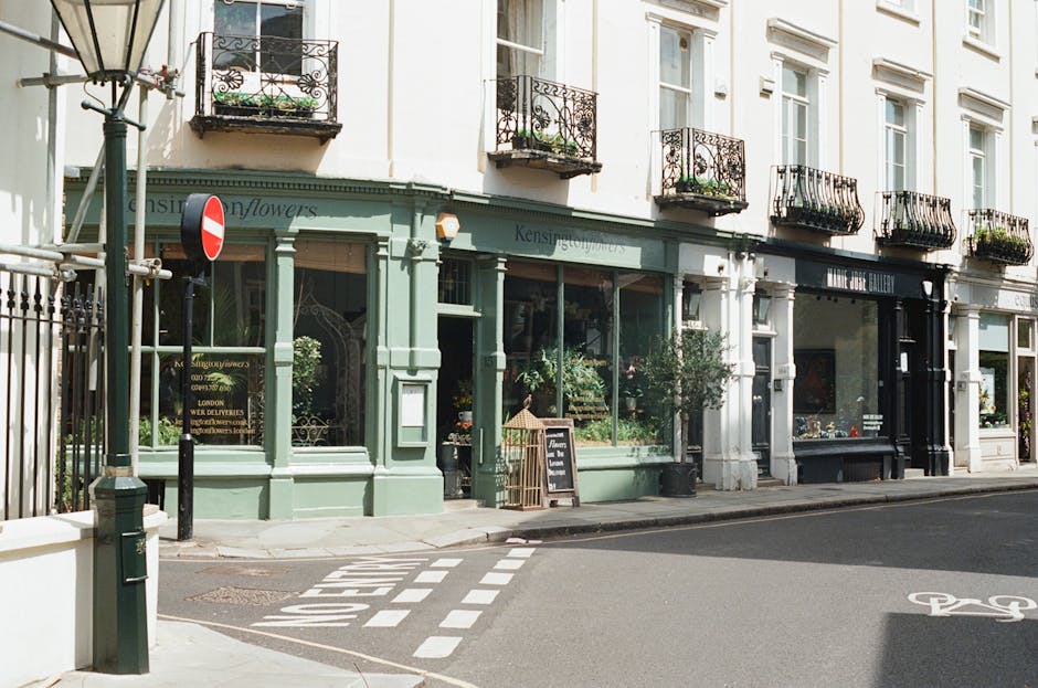 A florist shop named 'Kensington Flowers' with a pastel green exterior and large display windows showing floral arrangements inside. The shop is situated on a quiet street in Kensington, featuring a s