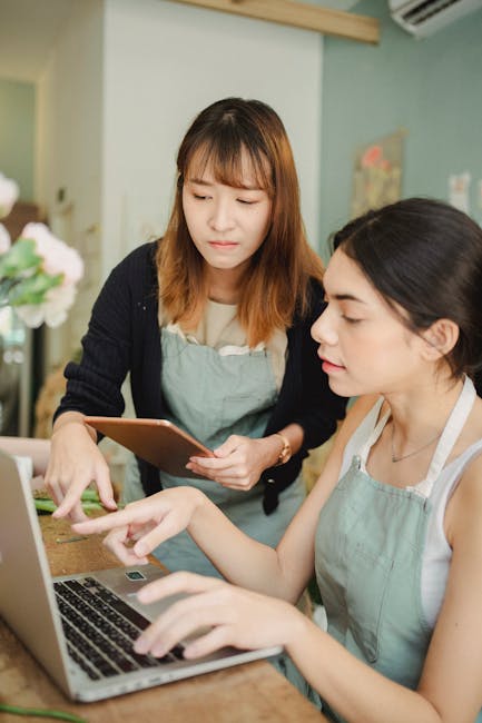 The image depicts two women working together in a flower shop workspace with pastel-colored walls, engaged in a discussion about floral arrangements. One woman with shoulder-length brown hair and wear
