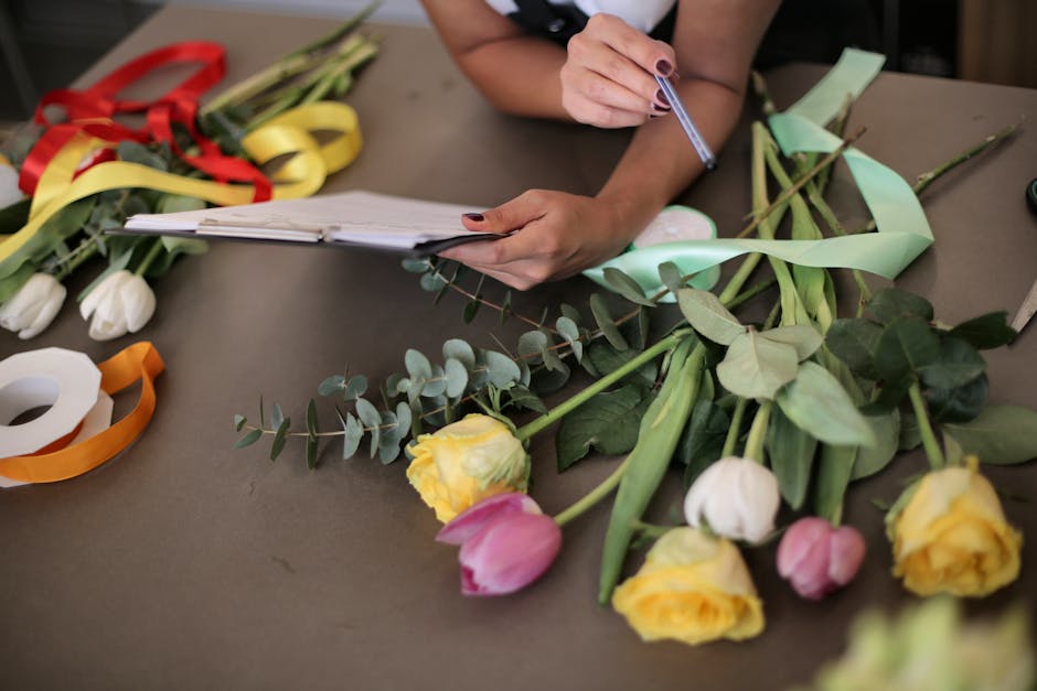 Close-up of a florist's workspace with a woman’s hand holding a pen and writing on a small notepad, surrounded by fresh wedding flower blooms including yellow, pink, and white tulips, accented with