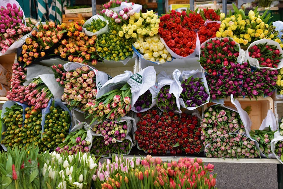A vibrant display of fresh flower bouquets at a flower market, featuring an array of tulips, roses, and daffodils in various colors including pink, white, red, purple, and yellow. The bouquets are wra