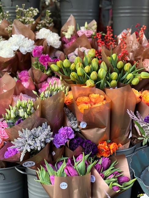 A vibrant display of freshly arranged flowers in brown paper wrapping, featuring a variety of blooms including white carnations, pink tulips, purple and lavender blossoms, orange roses, and green tuli