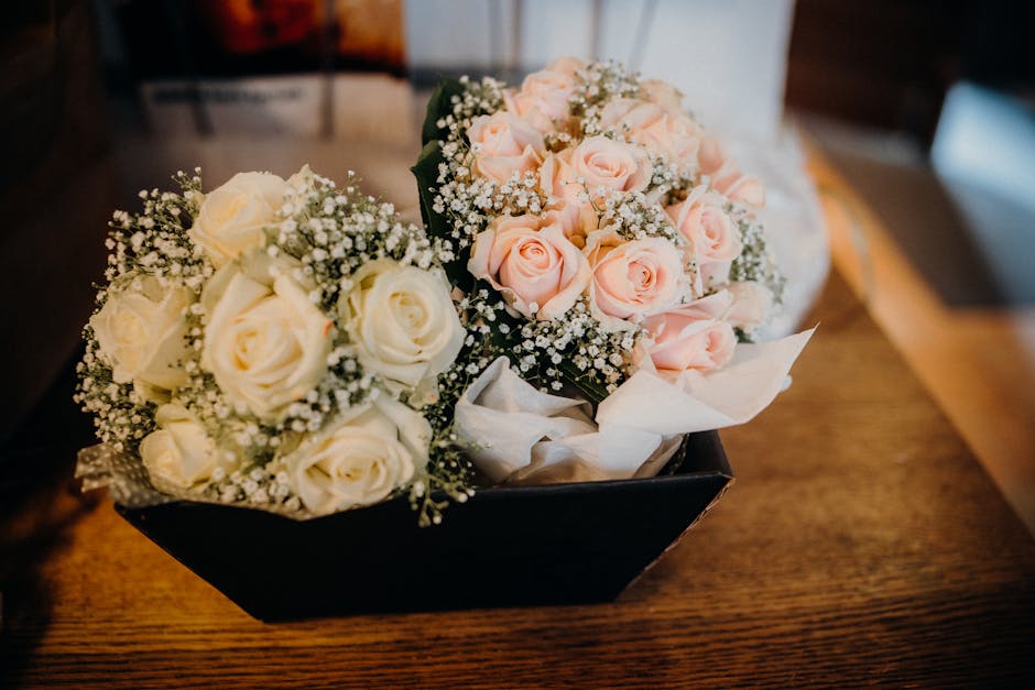 A floral arrangement featuring two bouquets of roses placed in a black rectangular box on a wooden surface. The bouquet on the left contains creamy white roses, accented with delicate sprigs of baby's
