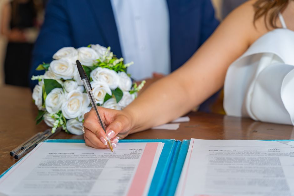 A floral arrangement featuring a bouquet of white roses with lush green foliage, placed on a wooden table beside a person signing a document with a silver and black pen. The bouquet is wrapped in clea