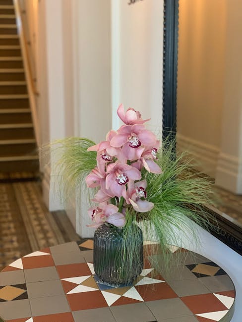 A tall, elegant floral arrangement featuring several large, pale pink orchids with ruffled petals and white centers, accented by wispy green foliage with fine, grass-like leaves. The orchids are place