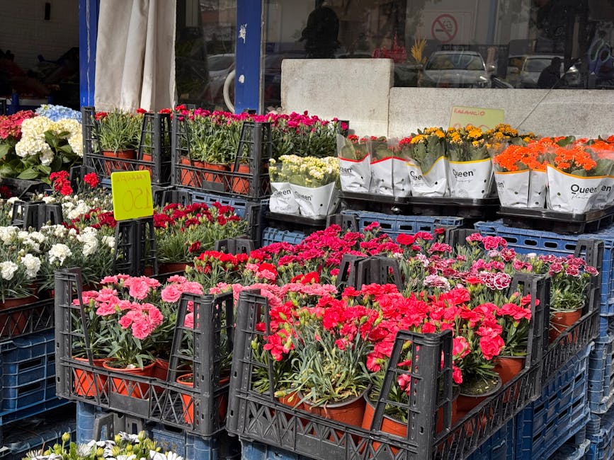 An outdoor display of various potted flowering plants arranged on black plastic crates at a garden centre. The flowers include pink, white, red, yellow, and orange blooms, with some wrapped in white p